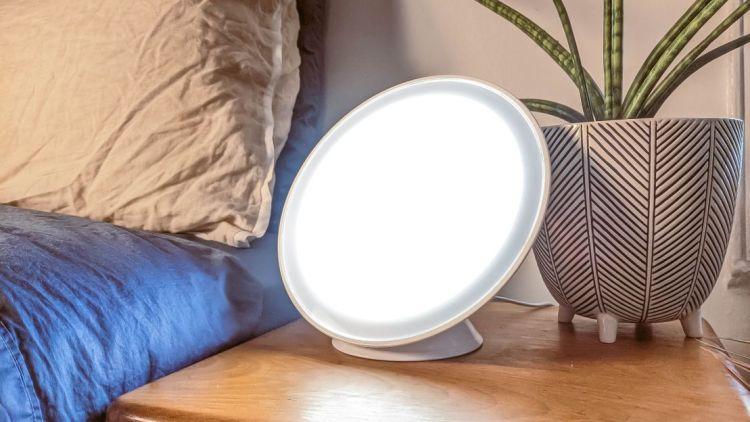 Light therapy lamp on a bedside table next to a pillow and a plant, used to improve mood and energy levels during dark winter months.