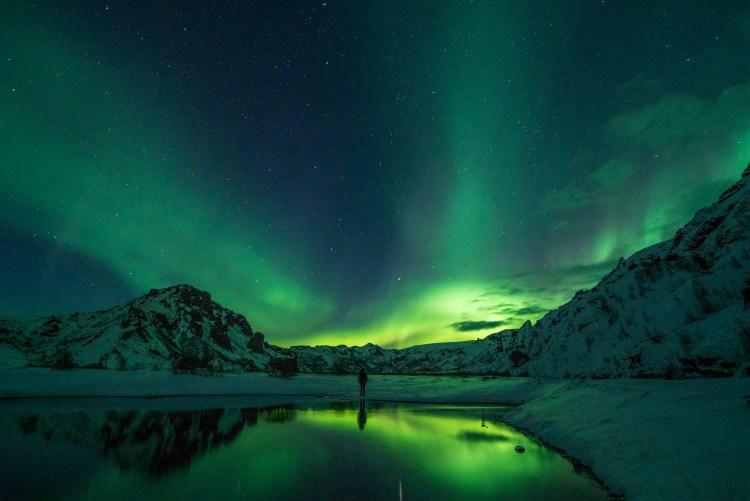 Bright green Northern Lights (Aurora Borealis) illuminating the snowy mountains and reflecting on a calm body of water, with a clear night sky in Iceland.
