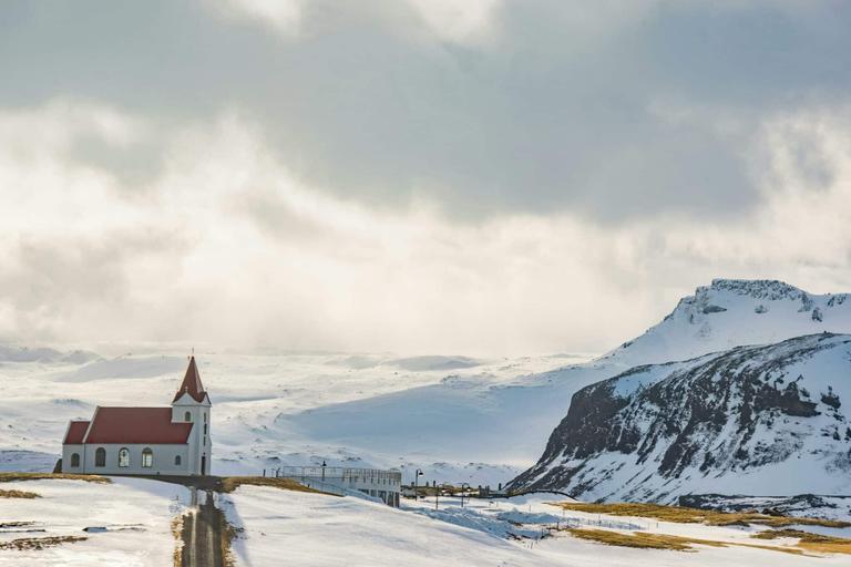 A small church with a red roof in a snowy, rural landscape in Iceland, with a cloudy sky and a distant mountain backdrop.
