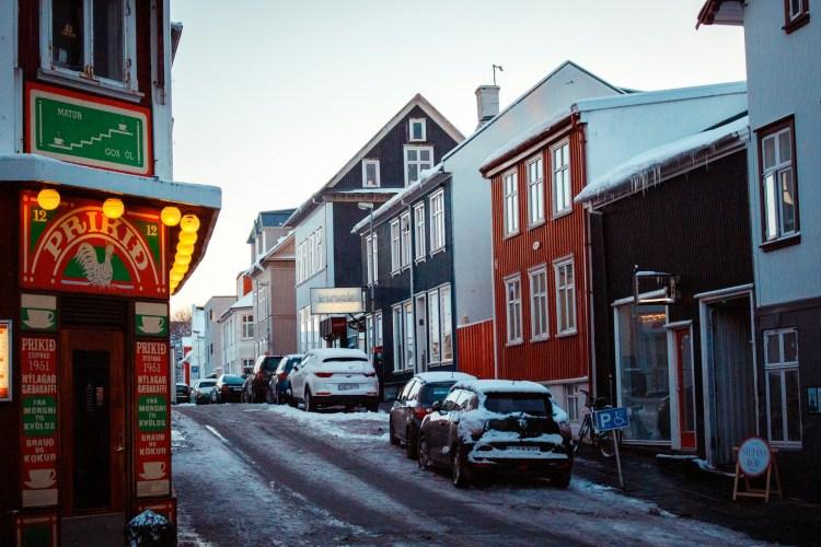 A snowy street in downtown Reykjavik, Iceland, with traditional corrugated iron houses and the Prikið bar, featuring colorful facades and cars covered in snow.